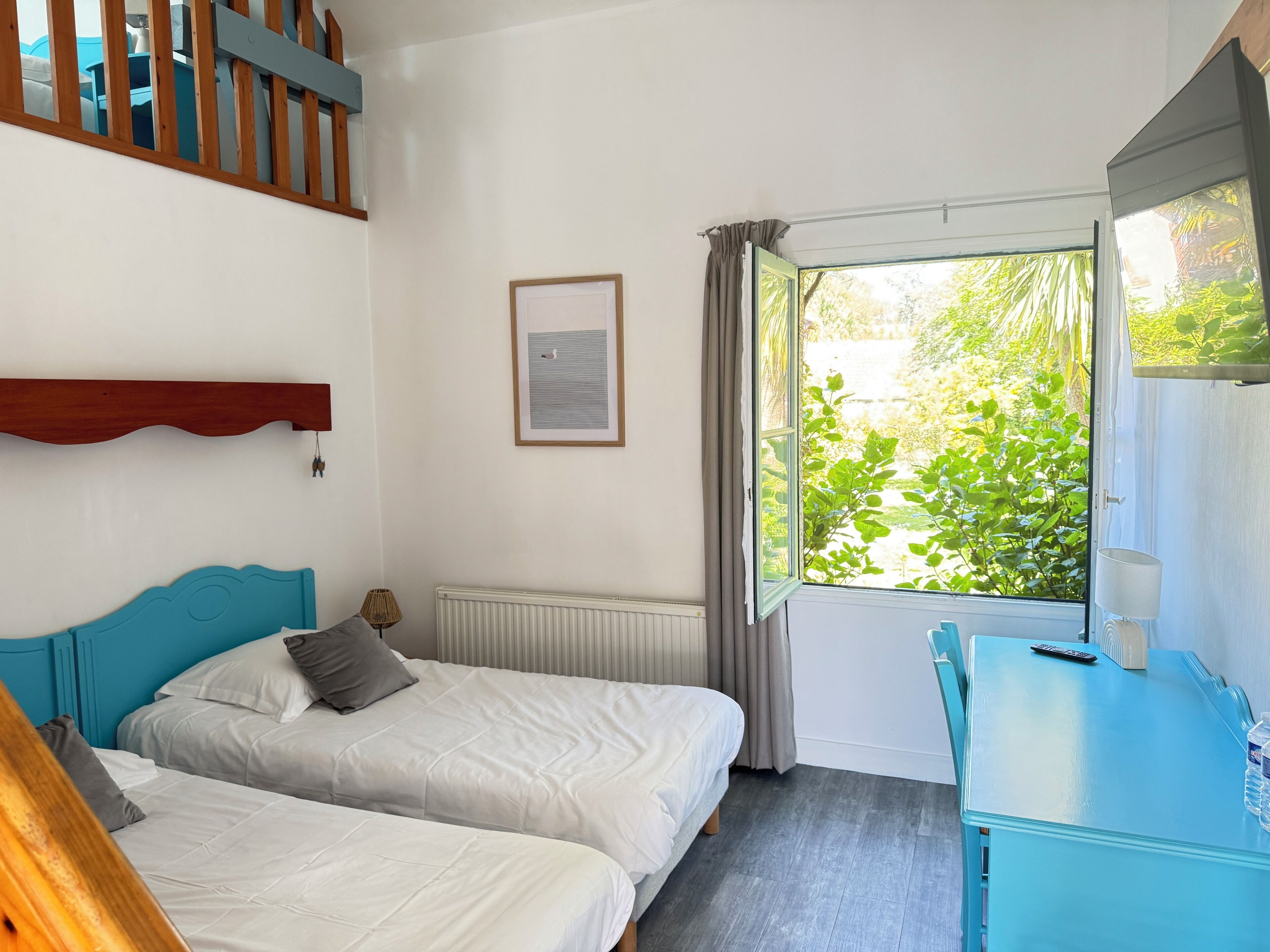 A hotel room with two beds, a desk, a window with curtains, a radiator, and a framed picture on the wall.