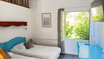 A hotel room with two beds, a desk, a window with curtains, a radiator, and a framed picture on the wall.