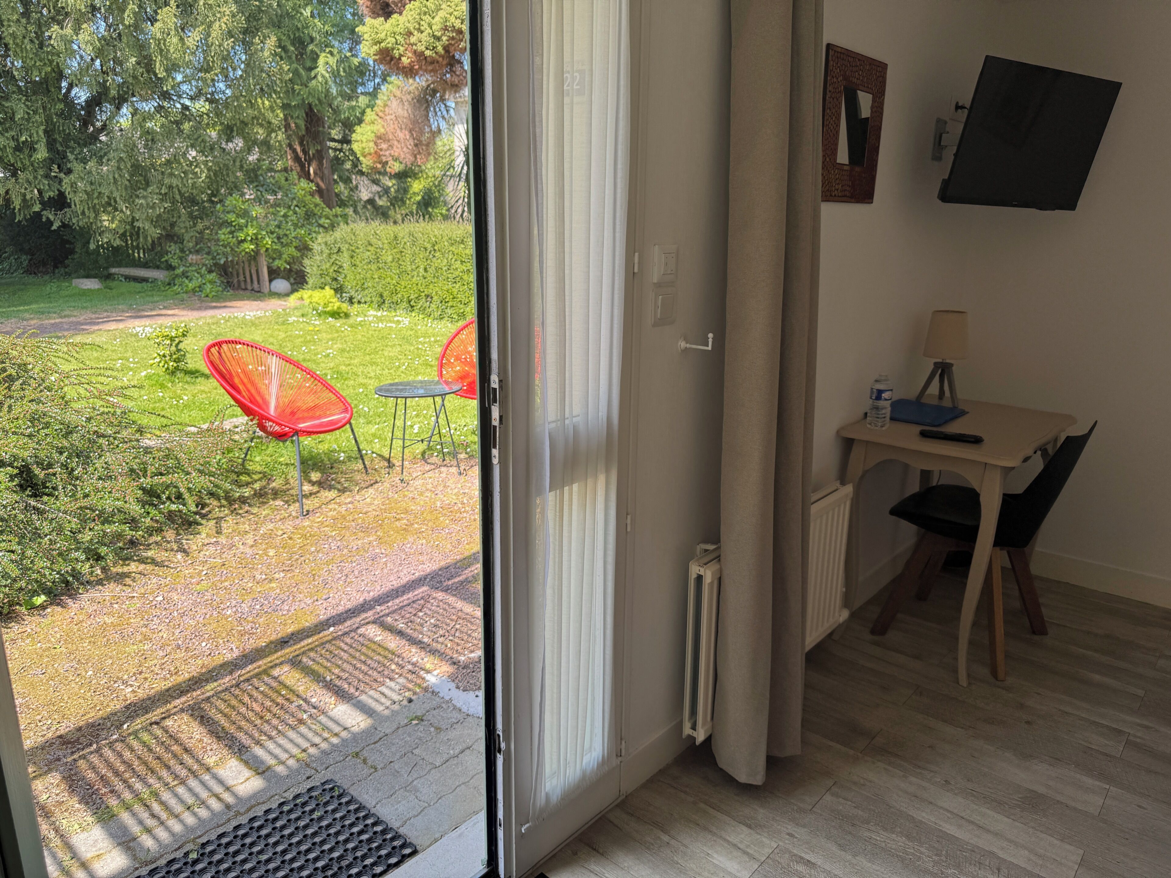 A room with a sliding glass door leading to a garden, a small table with a chair, a radiator, and a TV mounted on the wall.