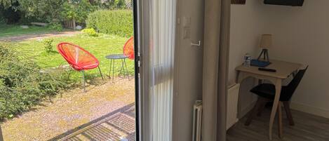 A room with a sliding glass door leading to a garden, a small table with a chair, a radiator, and a TV mounted on the wall.