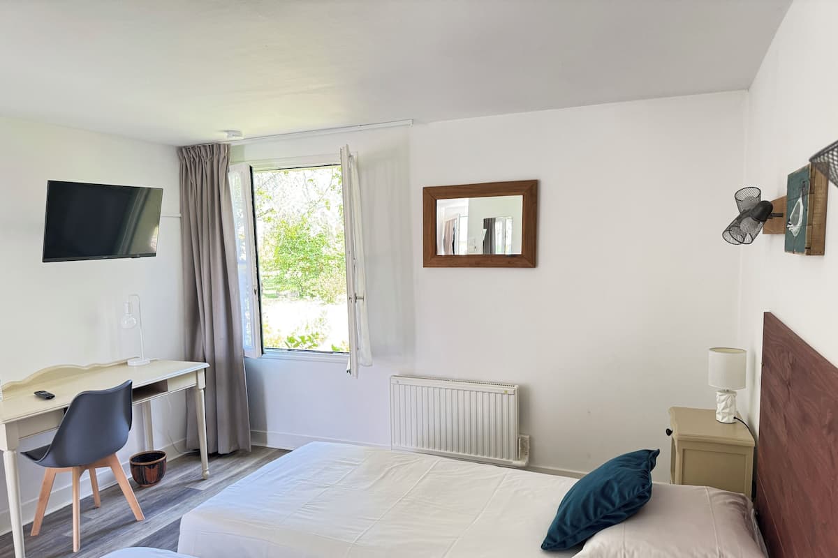Una habitación de hotel con una cama, un escritorio con una silla, televisión, ventana con cortinas, radiador y mesita de noche con lámpara.