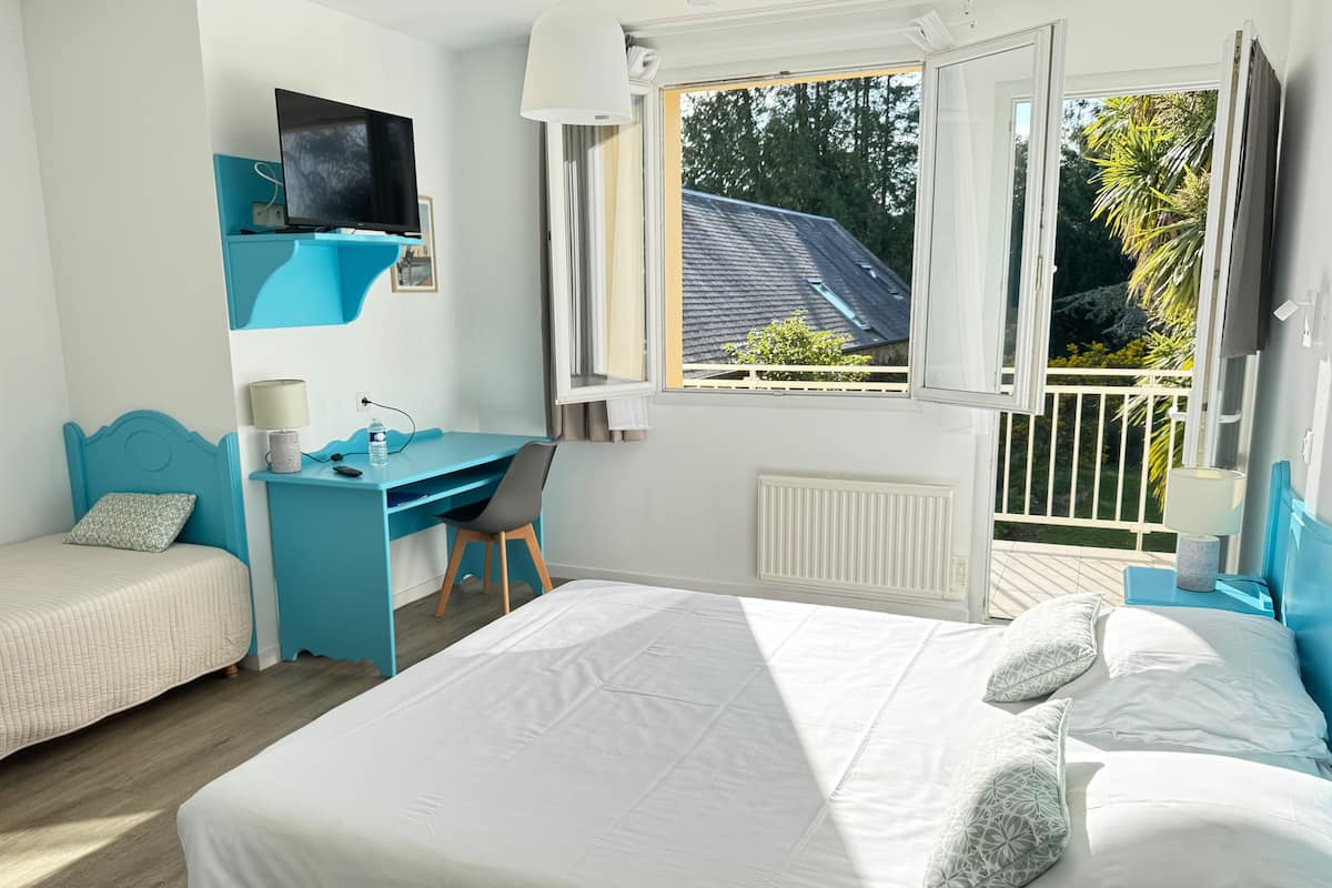 Un dormitorio con cama, escritorio, silla, televisor y ventana con vistas a árboles y una casa.