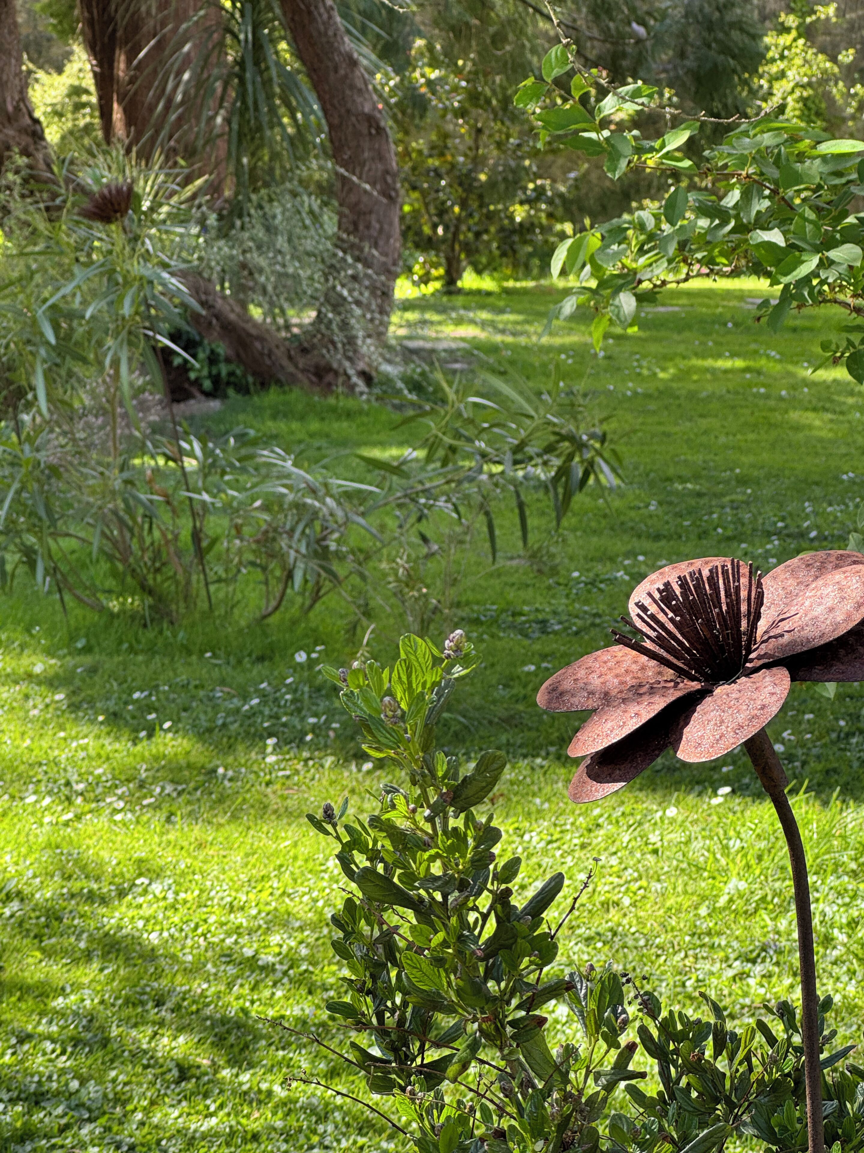 A garden with a prominent rusty metal flower in the foreground and various green plants and trees in the background.