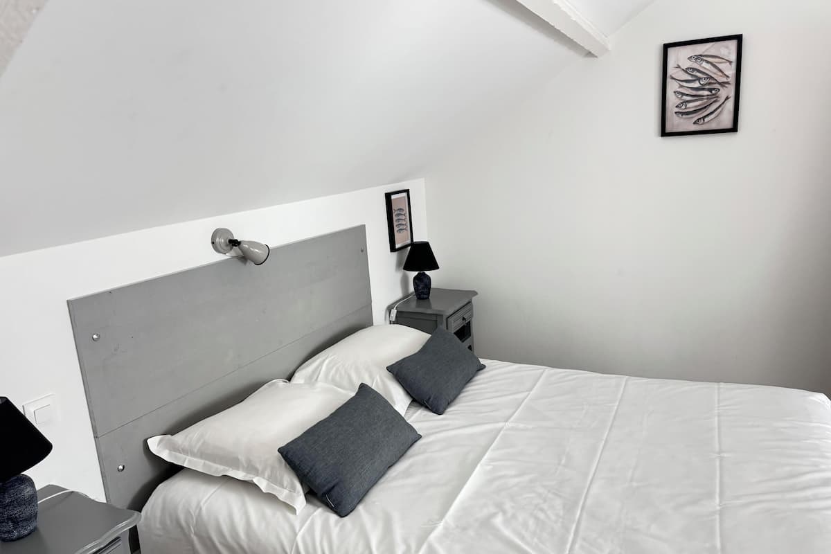 Un dormitorio moderno con cabecera gris, ropa de cama blanca, una mesita de noche con lámpara y una obra de arte enmarcada en la pared.