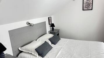 A modern bedroom with a grey headboard, white bedding, a bedside table with a lamp, and a framed artwork on the wall.