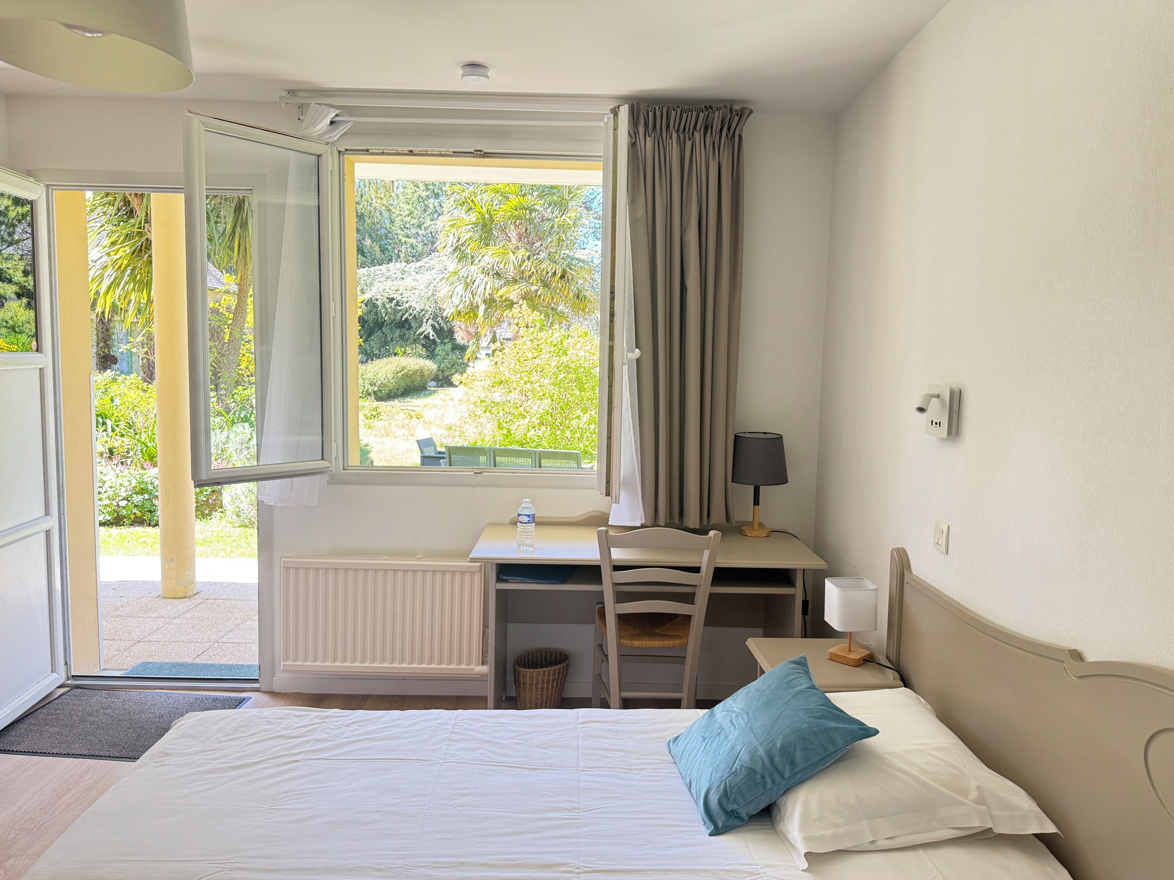 A bedroom with a bed, a desk with a chair, a window with curtains, and a view of greenery outside.