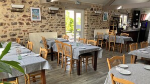 A restaurant with stone walls, multiple tables with grey tablecloths, wooden chairs, and a kitchen area visible in the background. - La Demeure Du Perron (Quettehou)