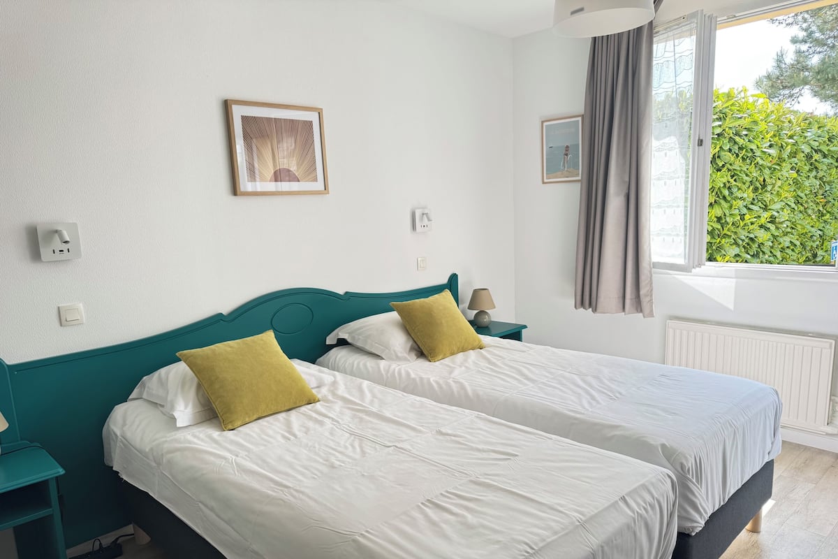 Una habitación de hotel con dos camas, una ventana con cortinas, un cuadro enmarcado en la pared y una mesita de noche con una lámpara.