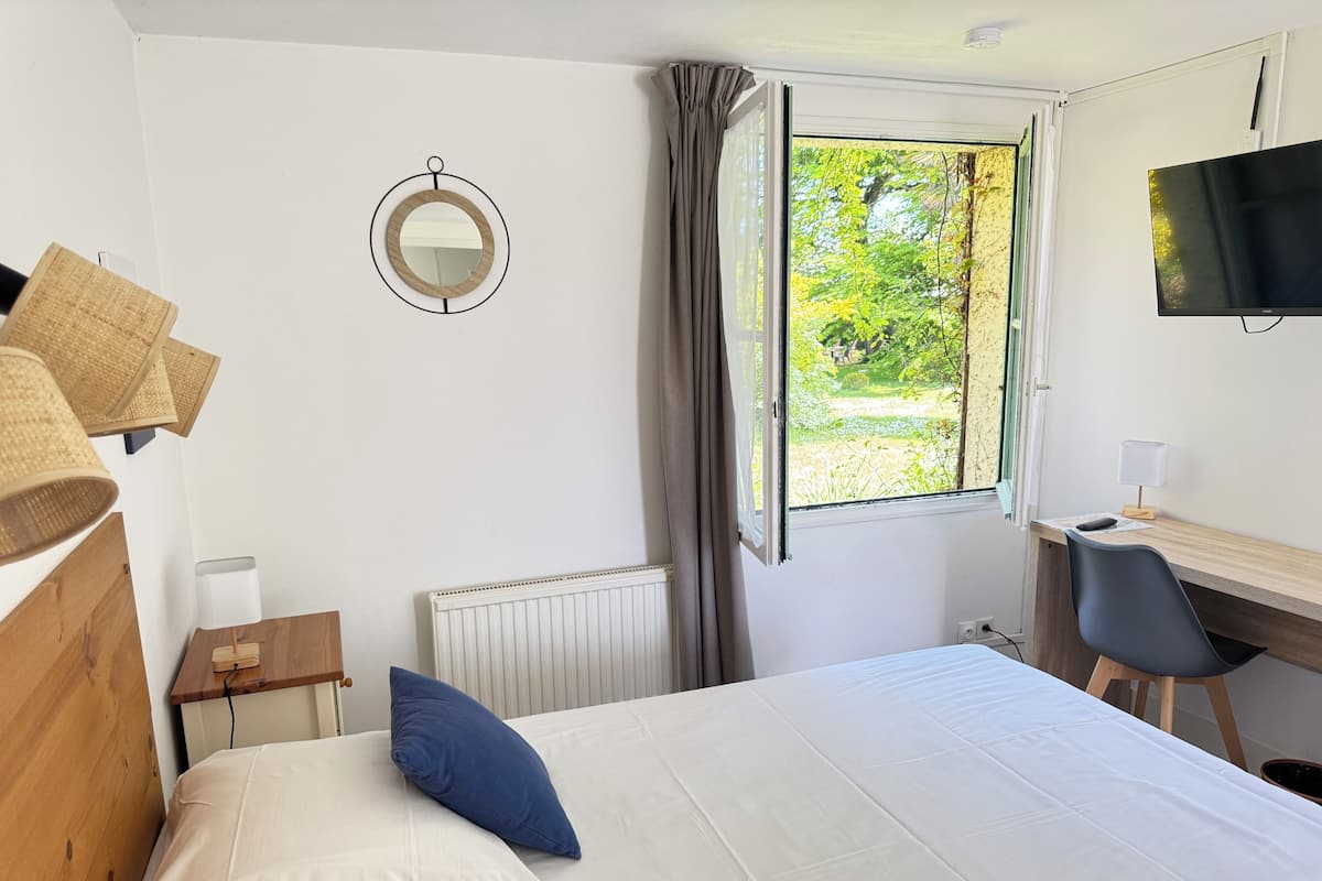 Un dormitorio con cama, escritorio, silla, televisor y ventana con vista a la vegetación.