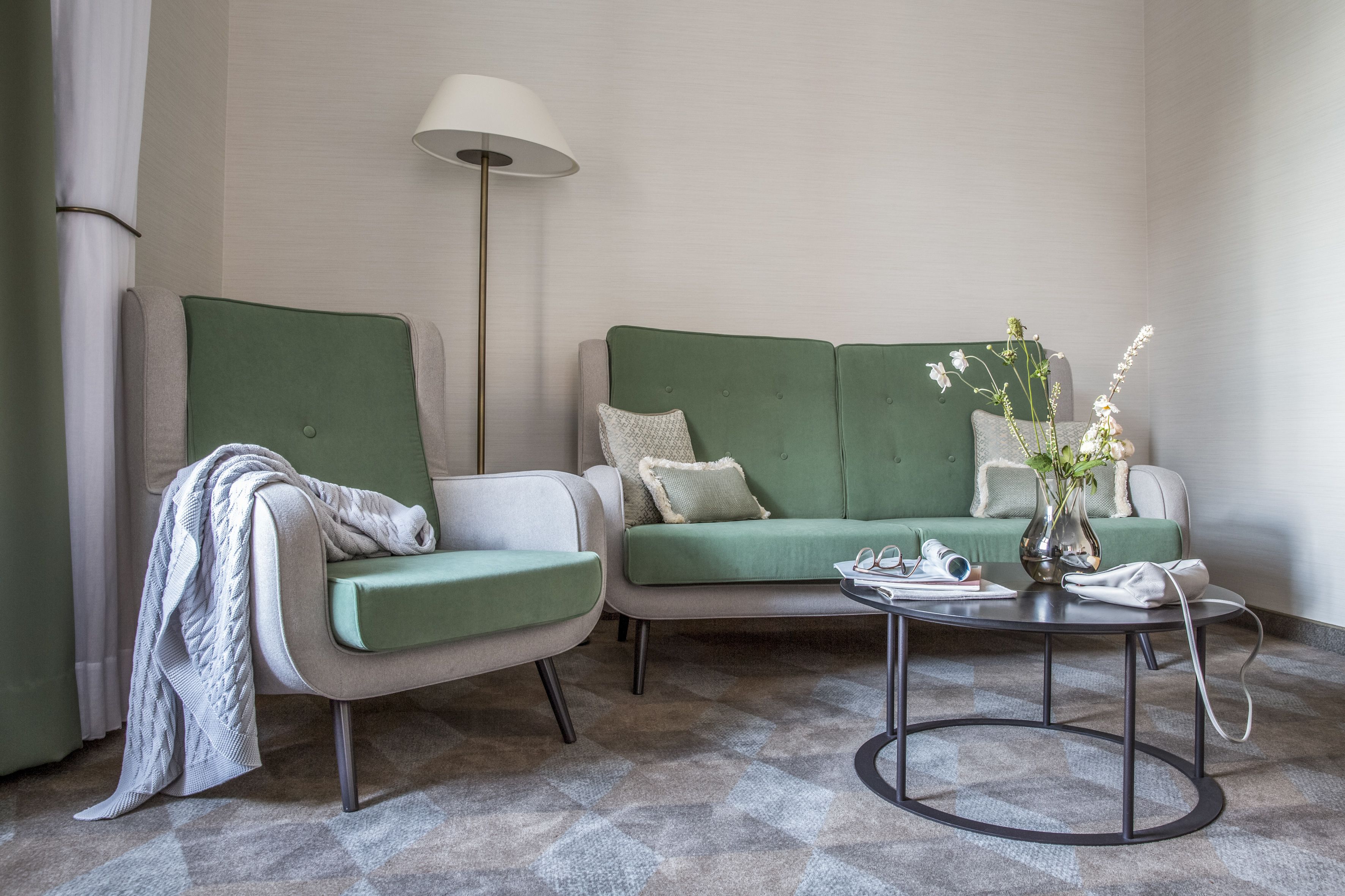 A modern hotel room with a green sofa, a matching armchair, a round coffee table, and a floor lamp.