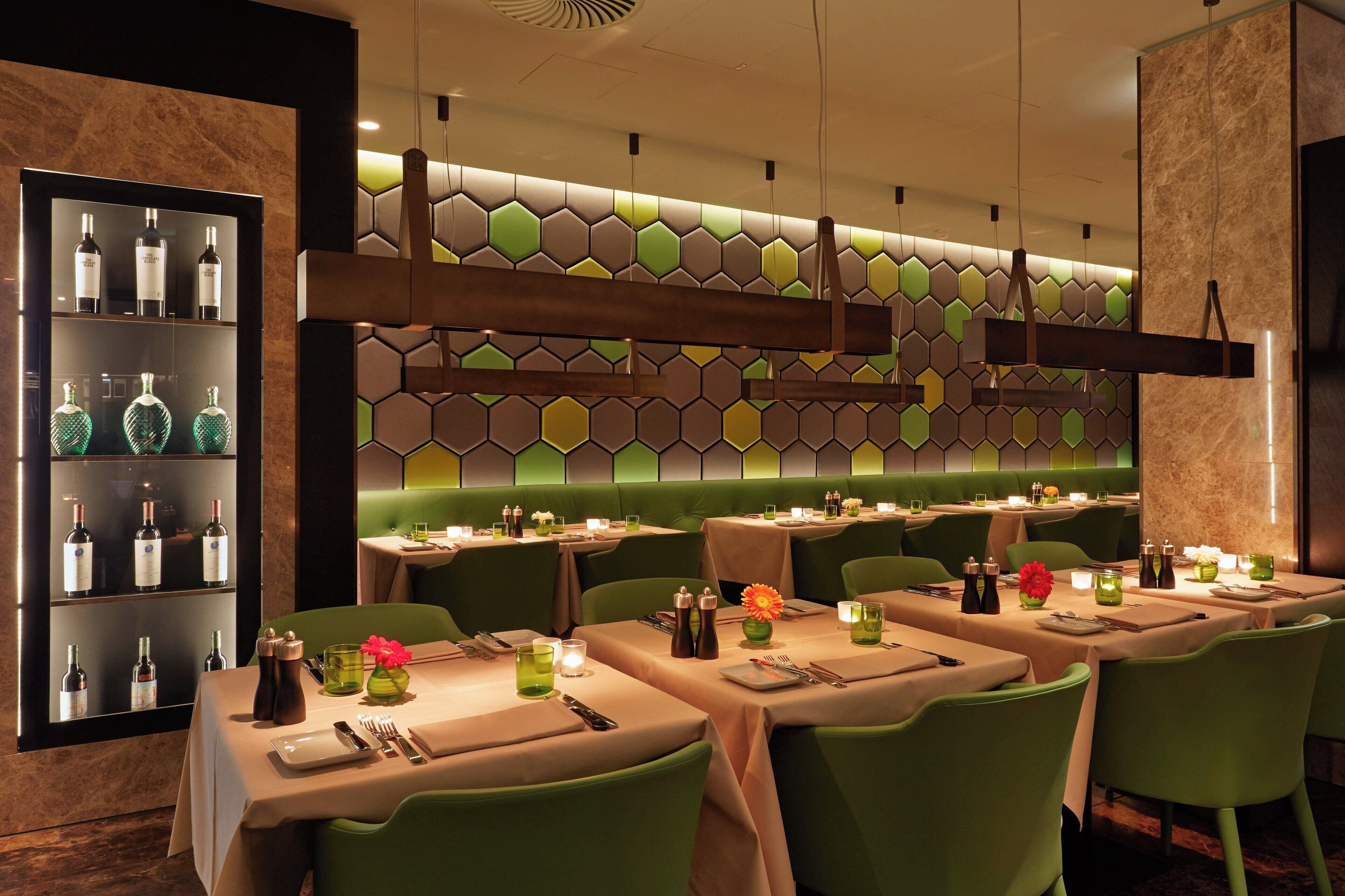 Un restaurante con asientos tipo banca verdes, mesas dispuestas con velas y una pared con diseño hexagonal.