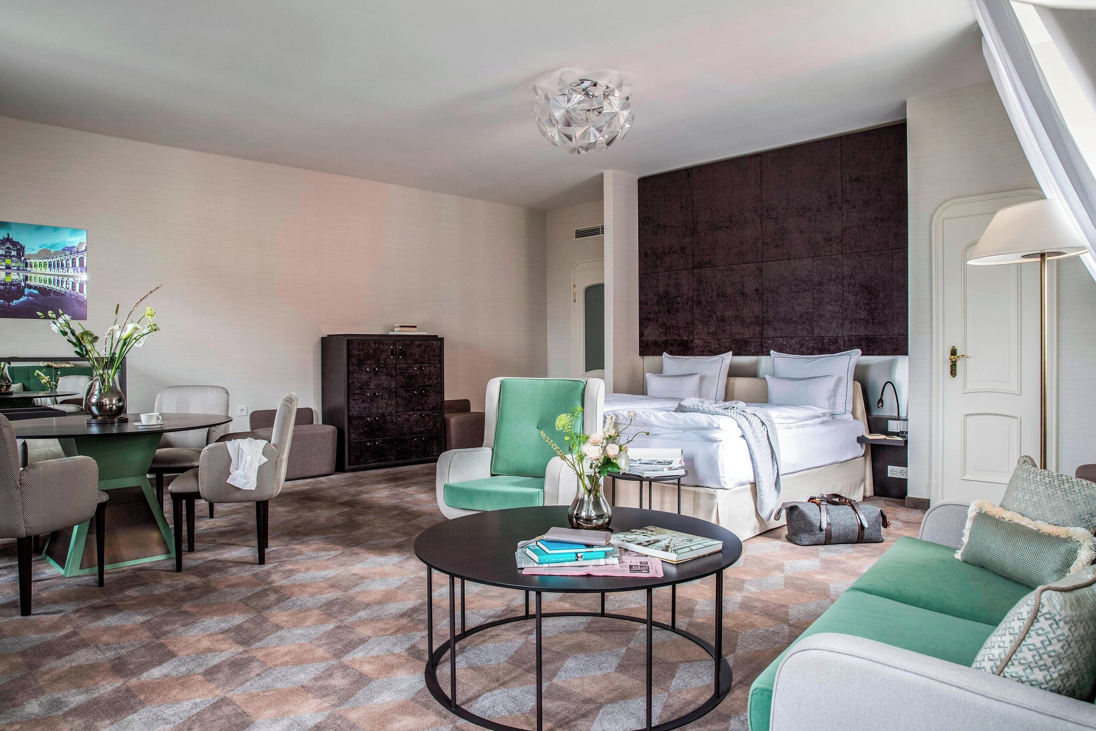 Une chambre d’hôtel avec un lit, un bureau, un fauteuil vert, une table basse ronde et un tableau au mur.