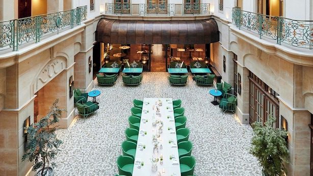 Un vaste atrium doté d’un haut plafond, une table centrale entourée de chaises et de nombreux balcons agrémentés de balustrades décoratives.