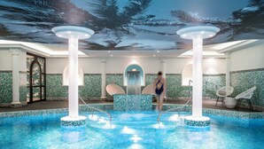 Une piscine intérieure ornée d’une fresque tropicale au plafond, d’une fontaine centrale et de colonnes blanches.