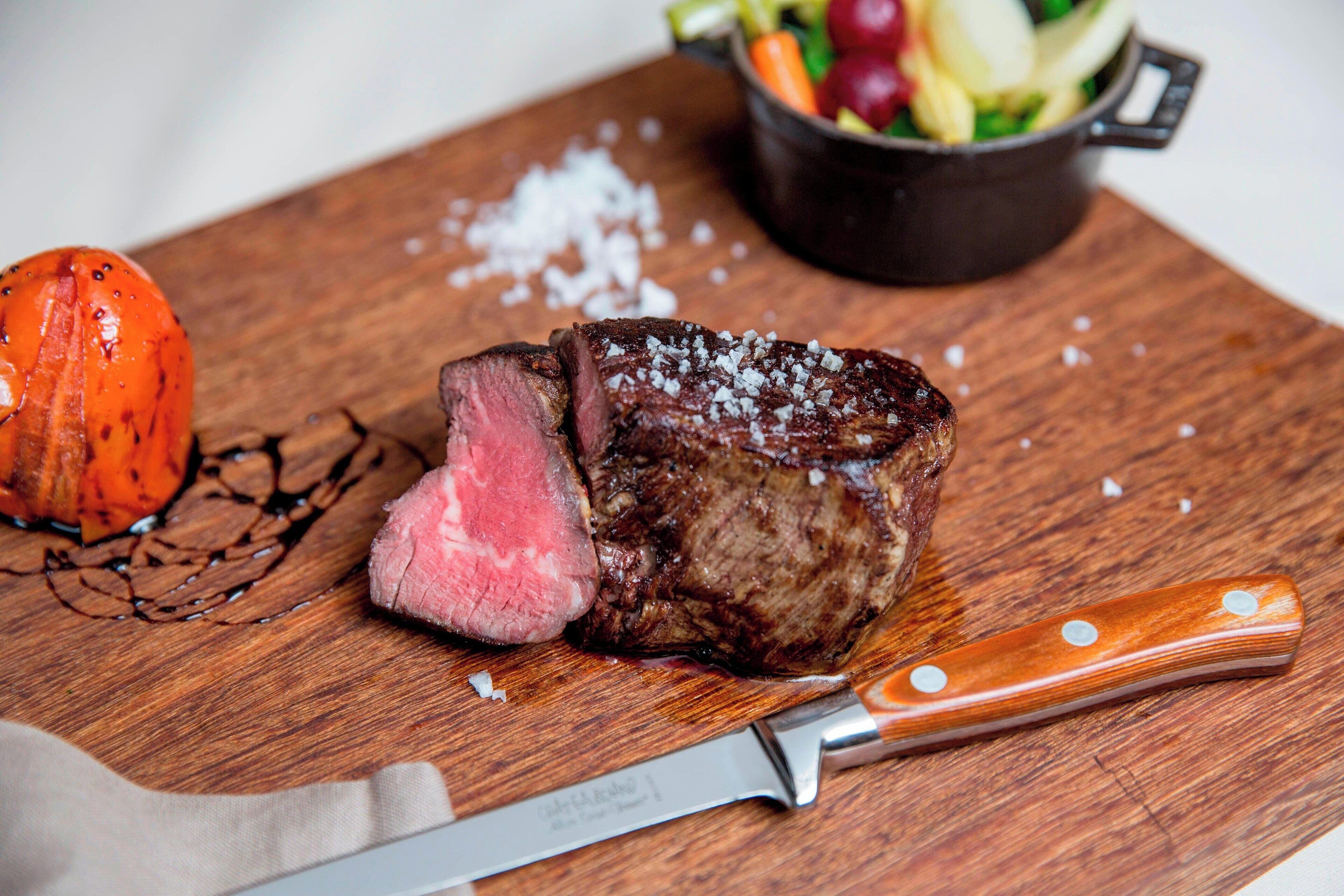 Un bistec sobre una tabla de madera con un cuchillo, un tazón de verduras y una olla al fondo.