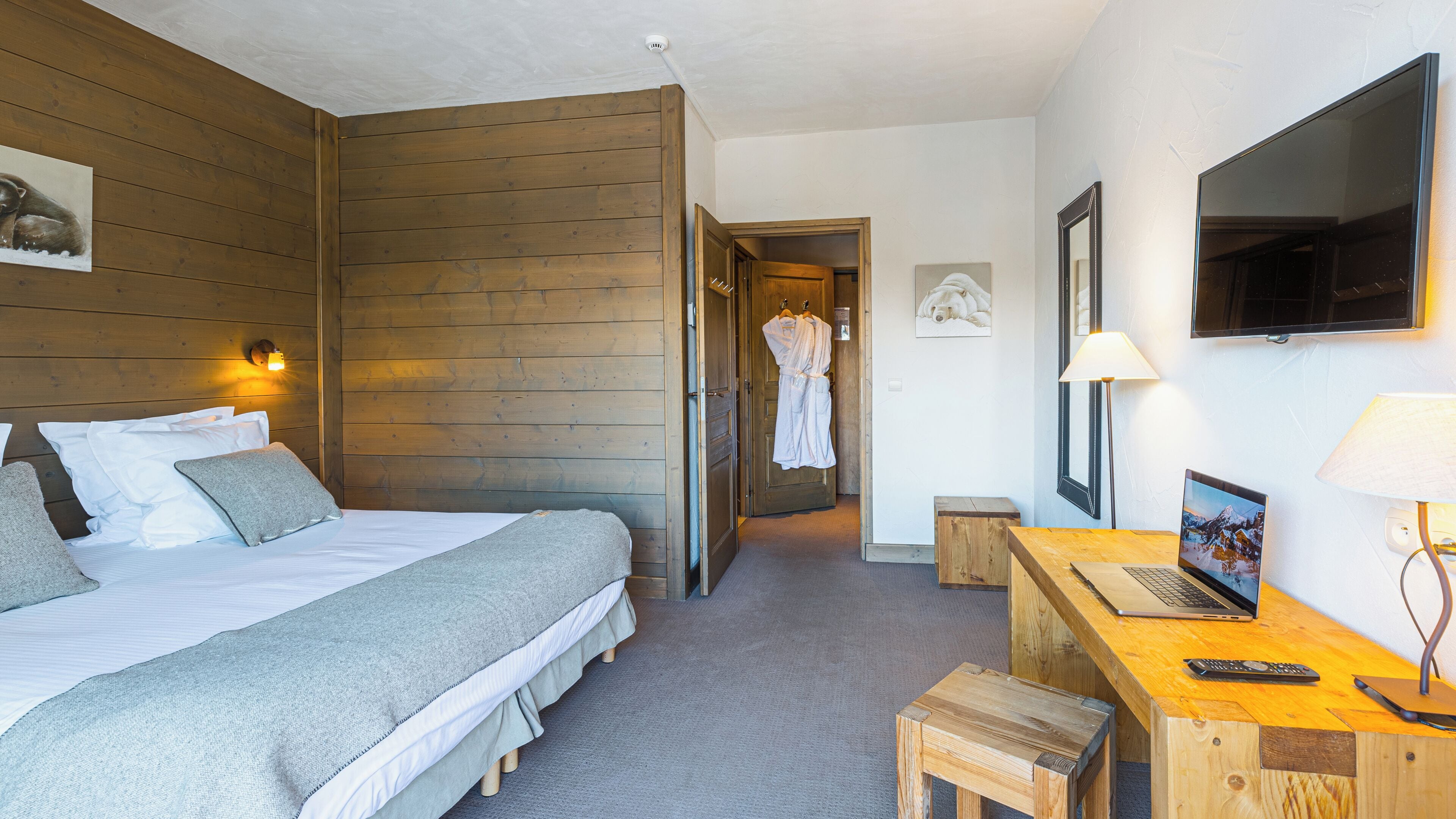 A hotel room with a wooden headboard, a bed with white bedding, a desk with a laptop, a TV, and a small stool.