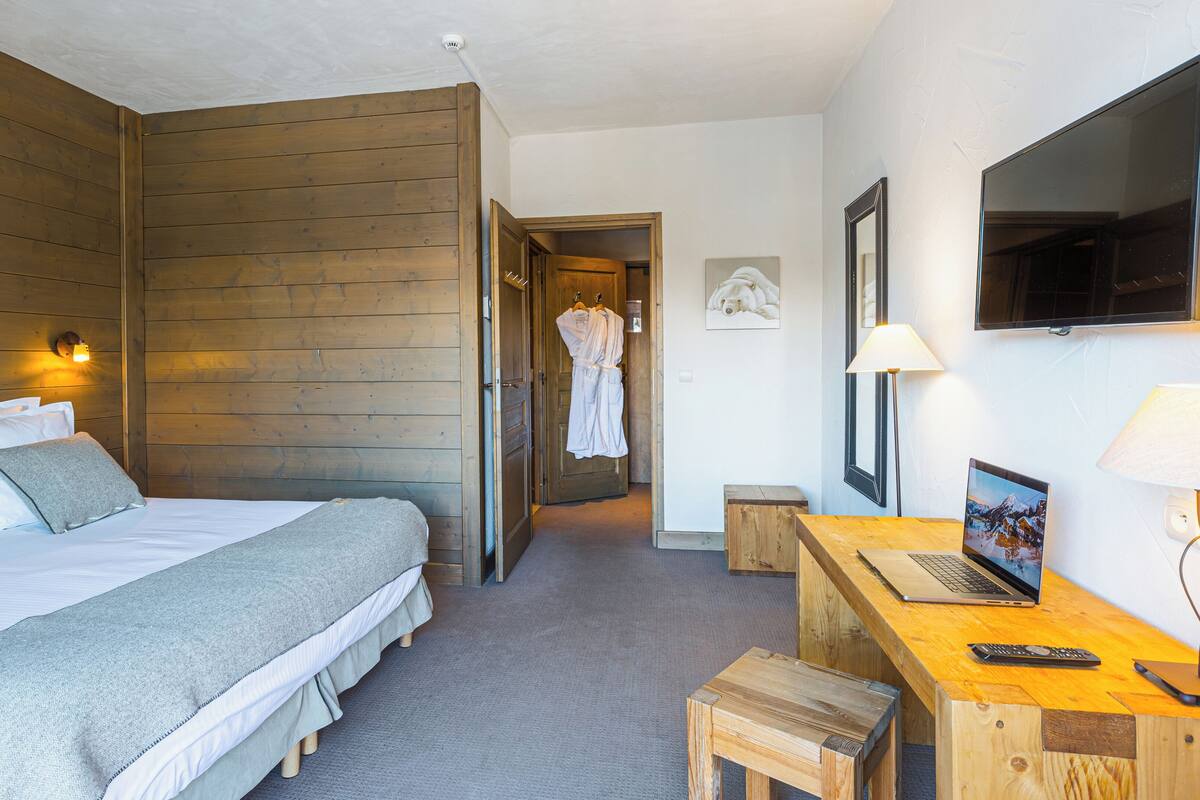 A hotel room with a wooden headboard, a bed with white bedding, a desk with a laptop, a TV, and a small stool.