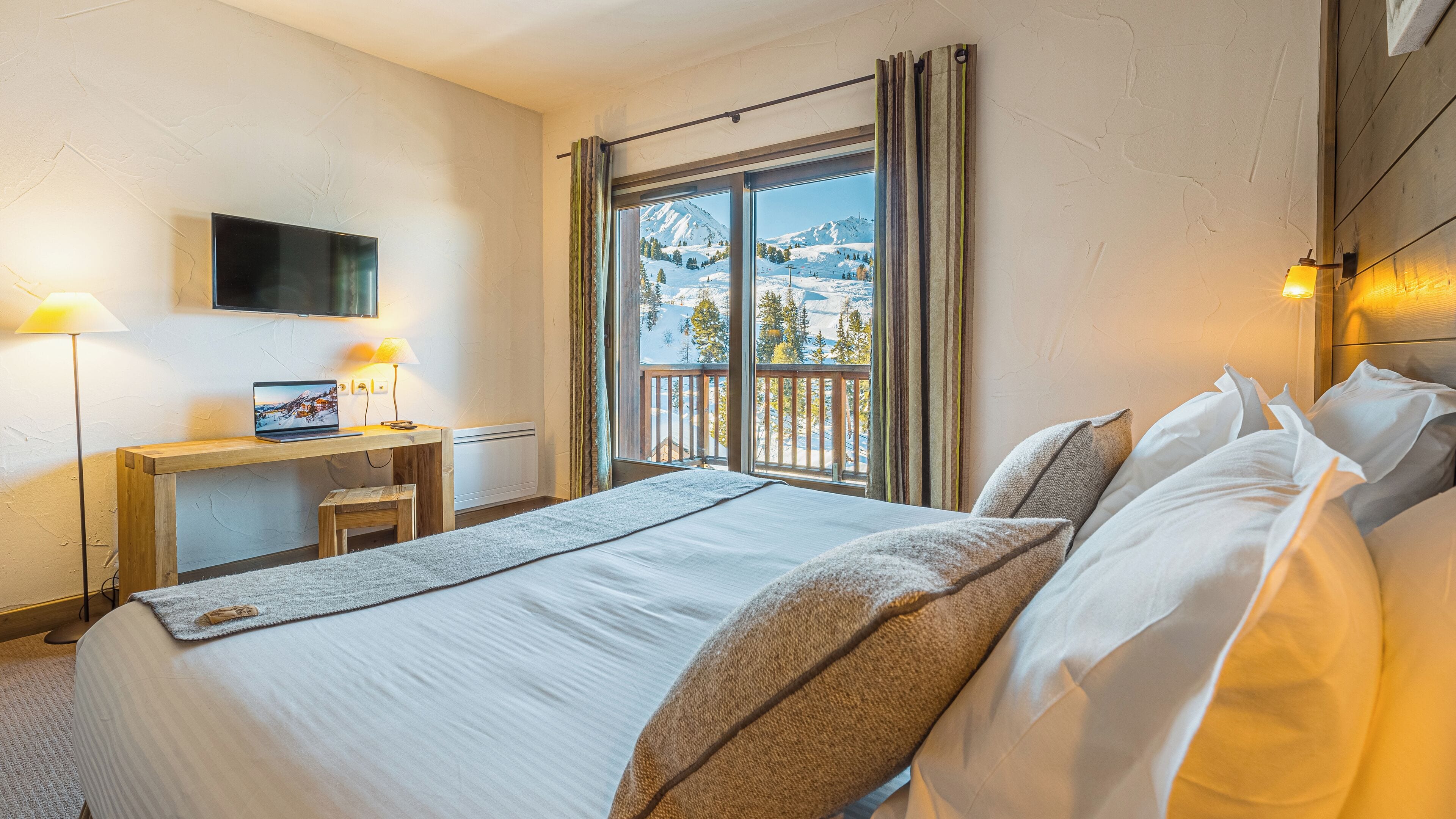 Una habitación de hotel con cama, escritorio con una computadora portátil, televisión y ventana con vista a la montaña.