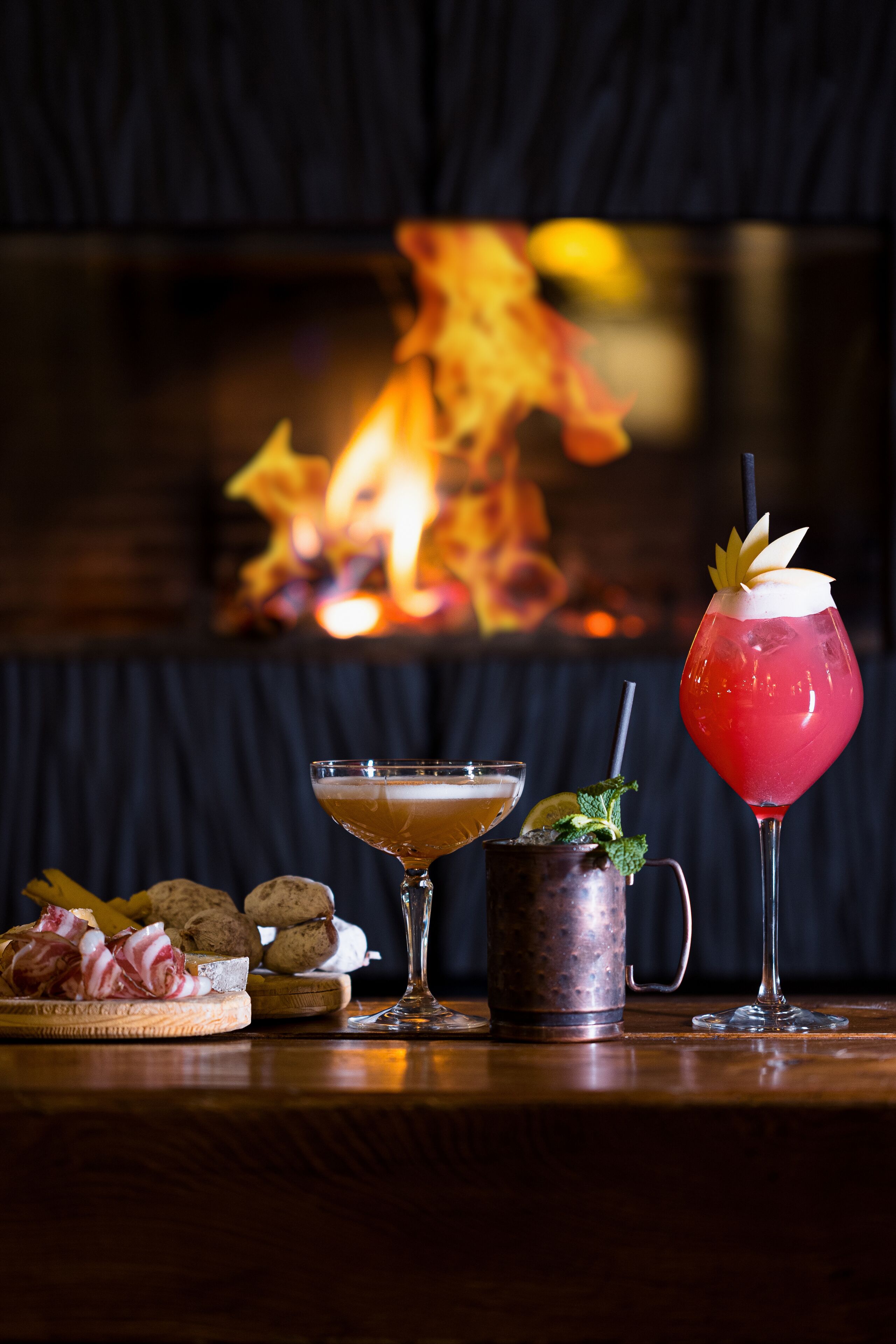 Eine Bar mit Kamin im Hintergrund, ein Martini-Glas, ein Cocktailglas mit Dekoration, ein Kupferbecher und eine Platte mit Snacks.