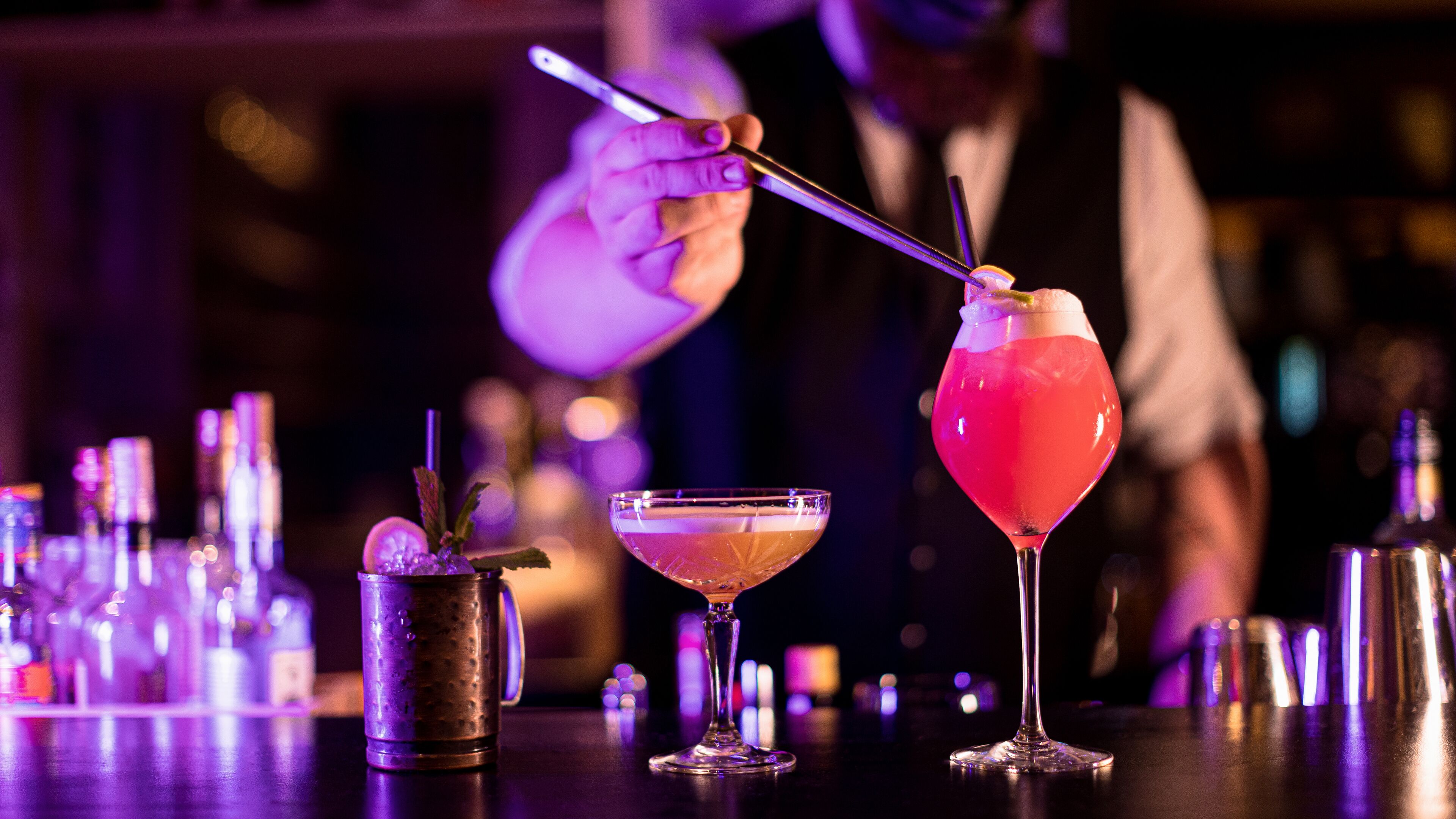 A bar setting with three cocktails, a bartender preparing drinks, and various bar tools.