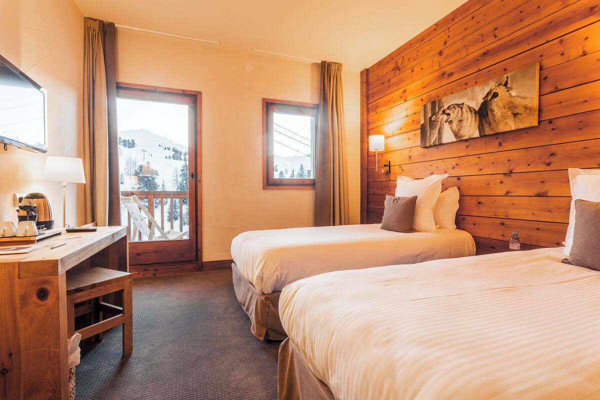 A hotel room with two beds, a wooden desk, a lamp, a painting, and a view of the mountains.