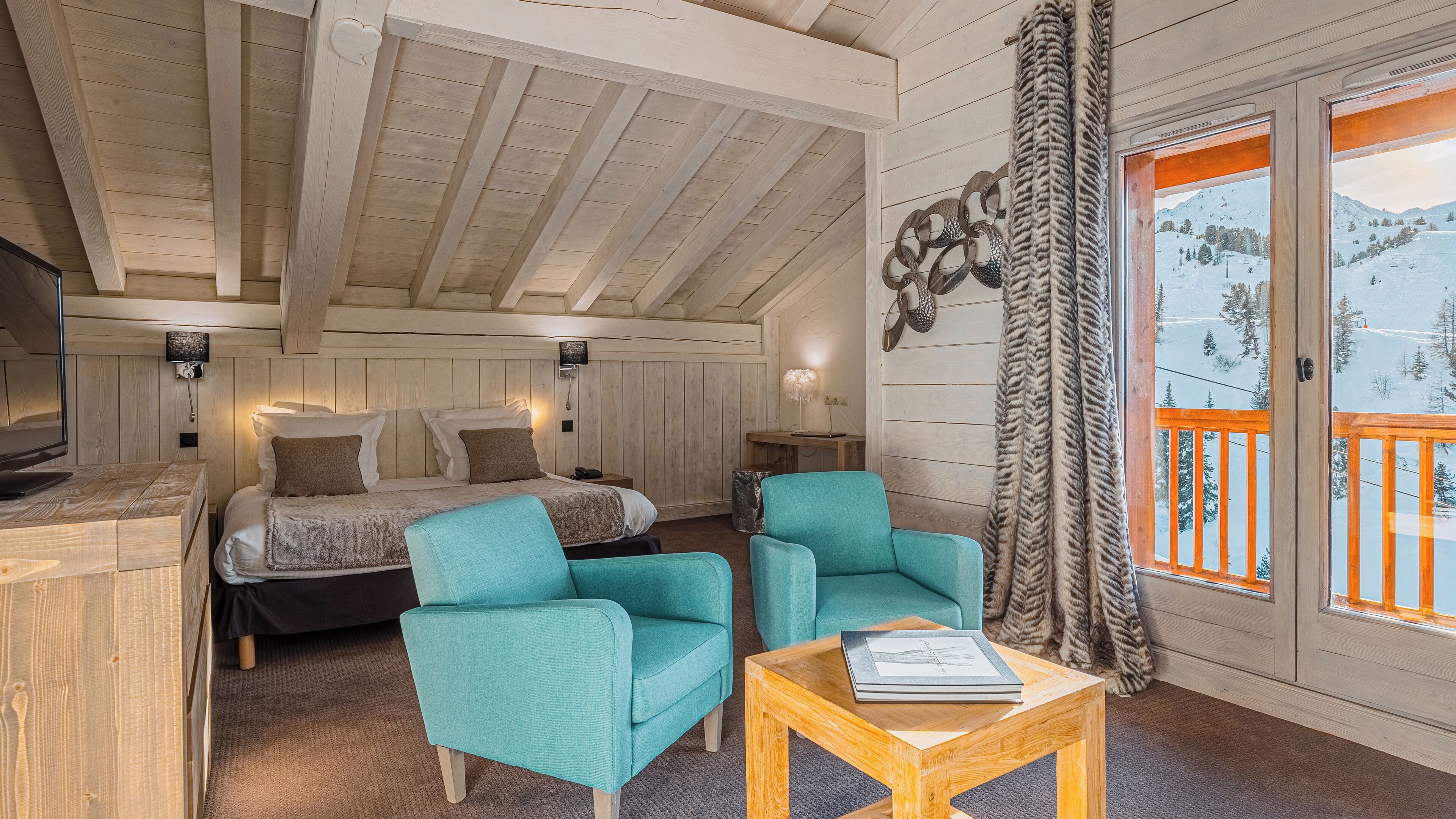A cozy room with a bed, two teal armchairs, a wooden coffee table, and a large window with a view of a snowy landscape.