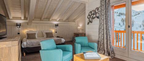 A cozy room with a bed, two teal armchairs, a wooden coffee table, and a large window with a view of a snowy landscape.