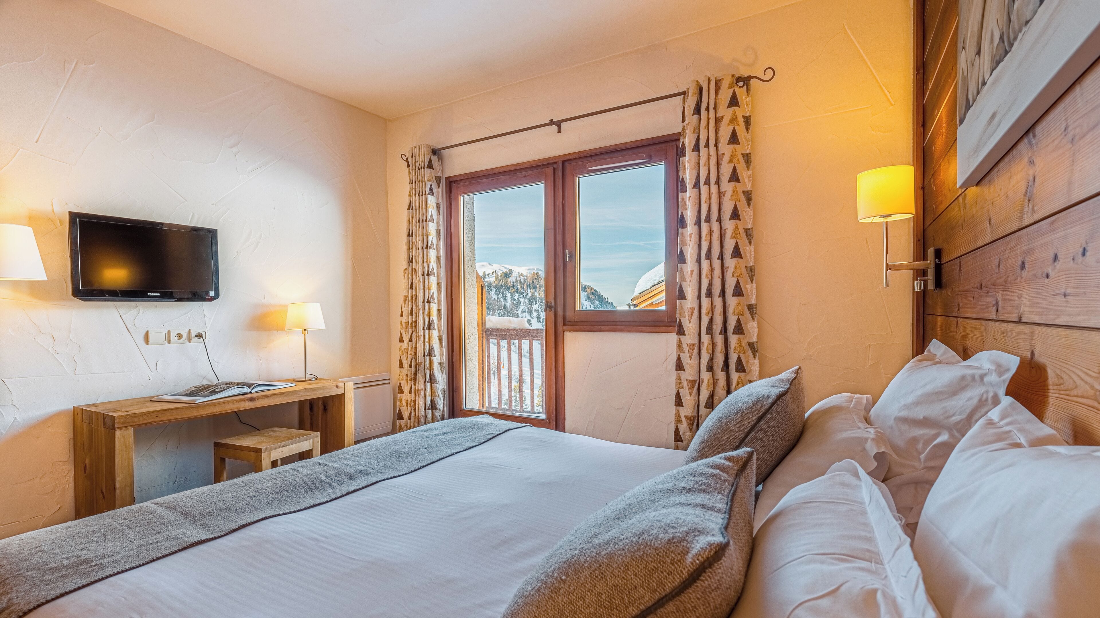 A hotel room with a bed, a desk with a lamp, a TV, and a window with curtains.