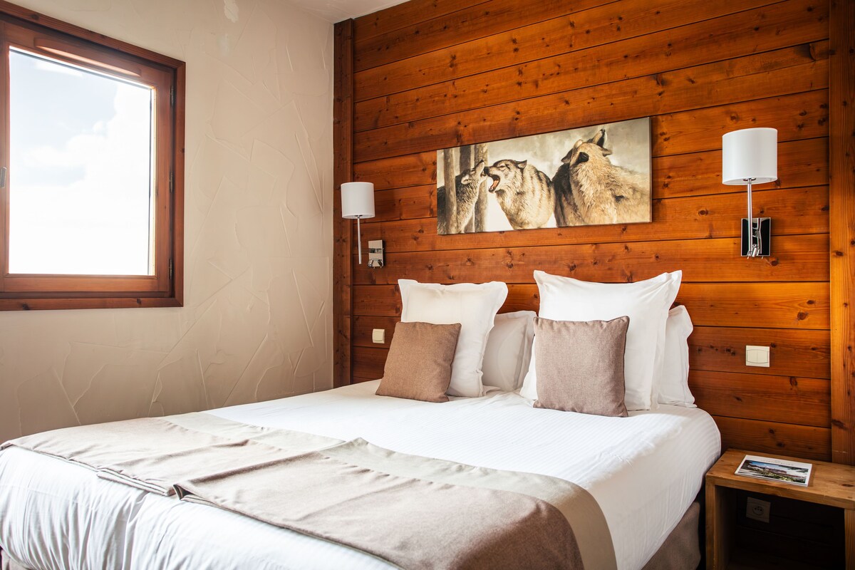A bedroom with a wooden headboard, a large window, two bedside lamps, and a painting of wolves.