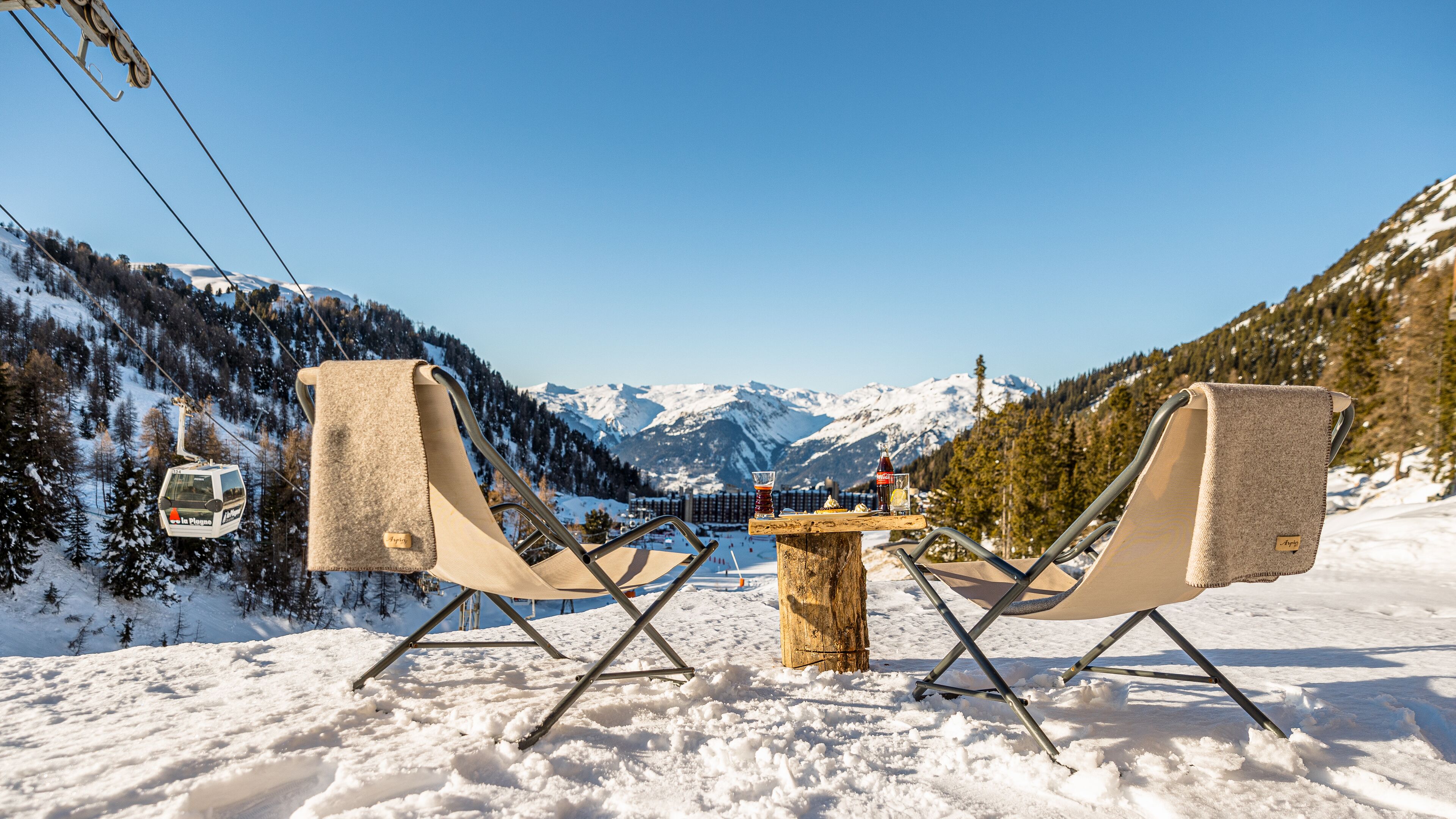 Un área de estar al aire libre cubierta de nieve con dos sillas y una mesa, con vistas a una cordillera y un telesilla.
