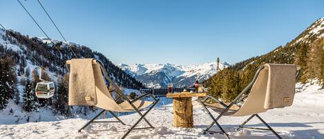 Un área de estar al aire libre cubierta de nieve con dos sillas y una mesa, con vistas a una cordillera y un telesilla.