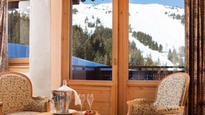 Une pièce chaleureuse au décor intérieur en bois, avec une grande fenêtre donnant sur une montagne enneigée, deux fauteuils rembourrés, une petite table ronde avec une théière, des verres et un bol de fraises.