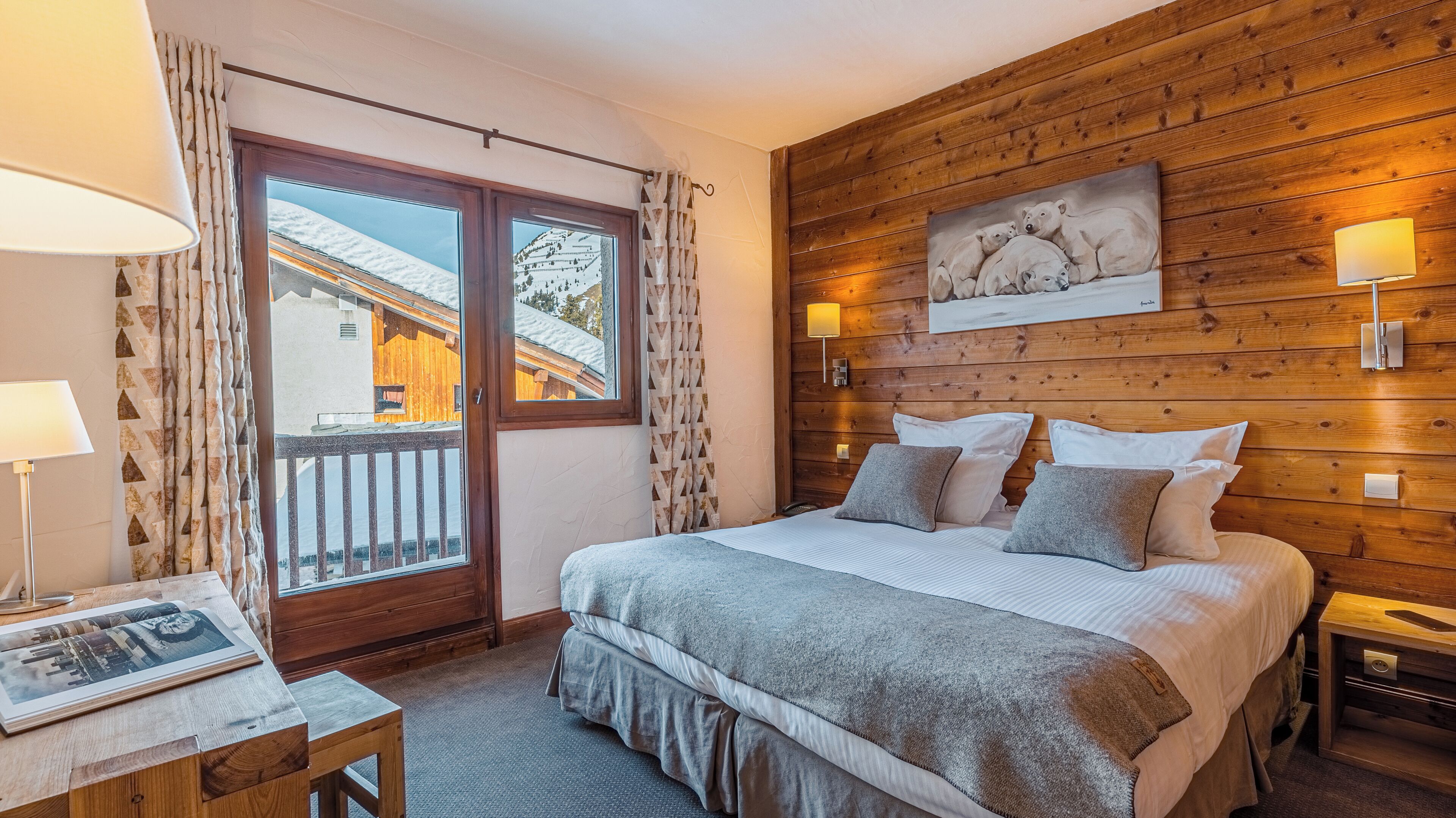 A bedroom with a wooden headboard, a large bed with pillows, a bedside table with a lamp, and a view of a snowy landscape through a sliding door.