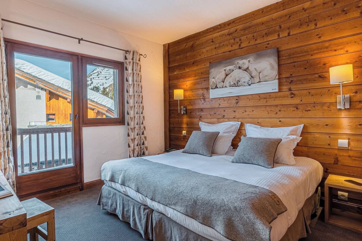 A bedroom with a wooden headboard, a large bed with pillows, a bedside table with a lamp, and a view of a snowy landscape through a sliding door.