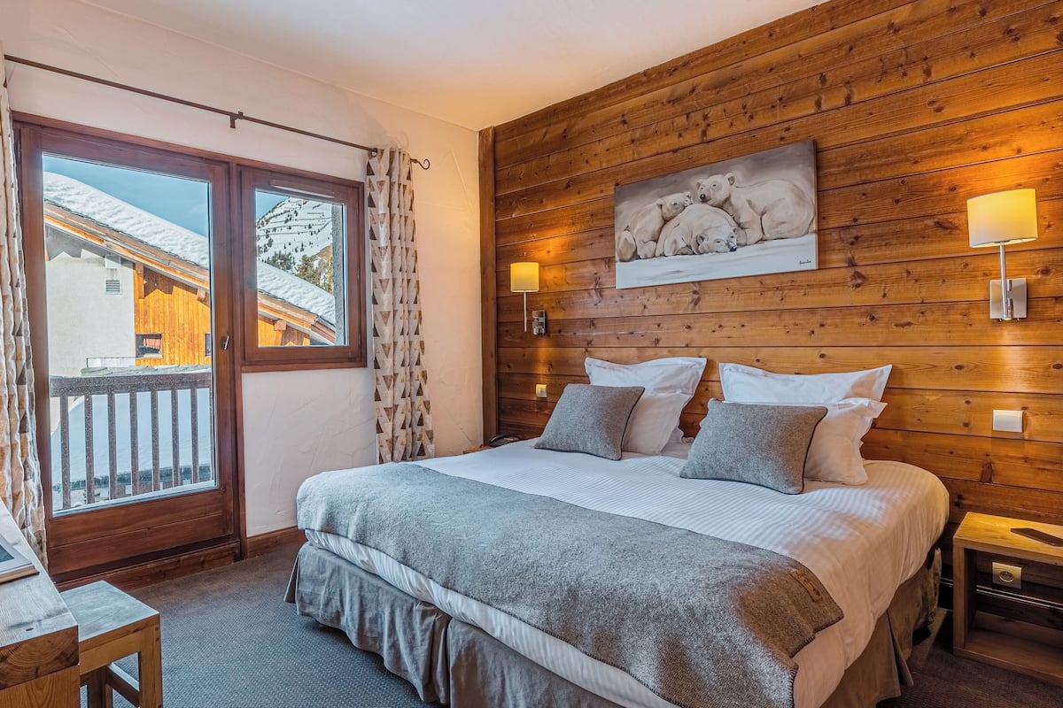 A bedroom with a wooden headboard, a large bed with pillows, a bedside table with a lamp, and a view of a snowy landscape through a sliding door.