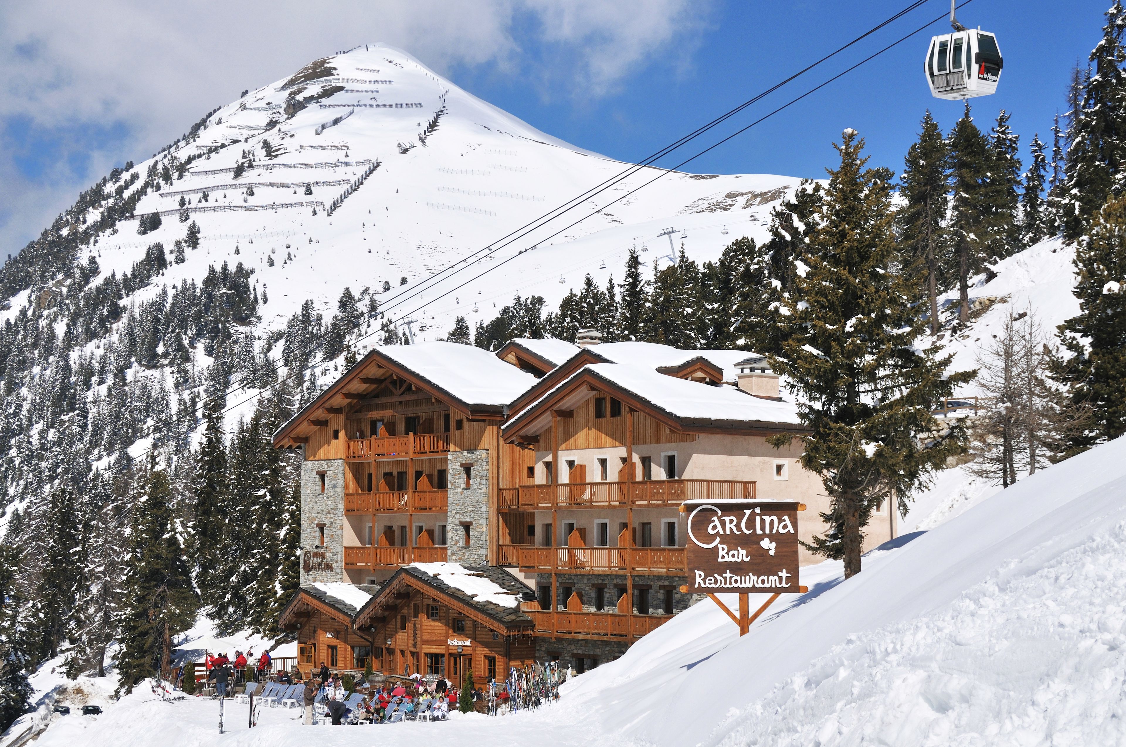A large wooden building with a sign reading "Carina Bar Restaurant" in a snowy mountainous area.