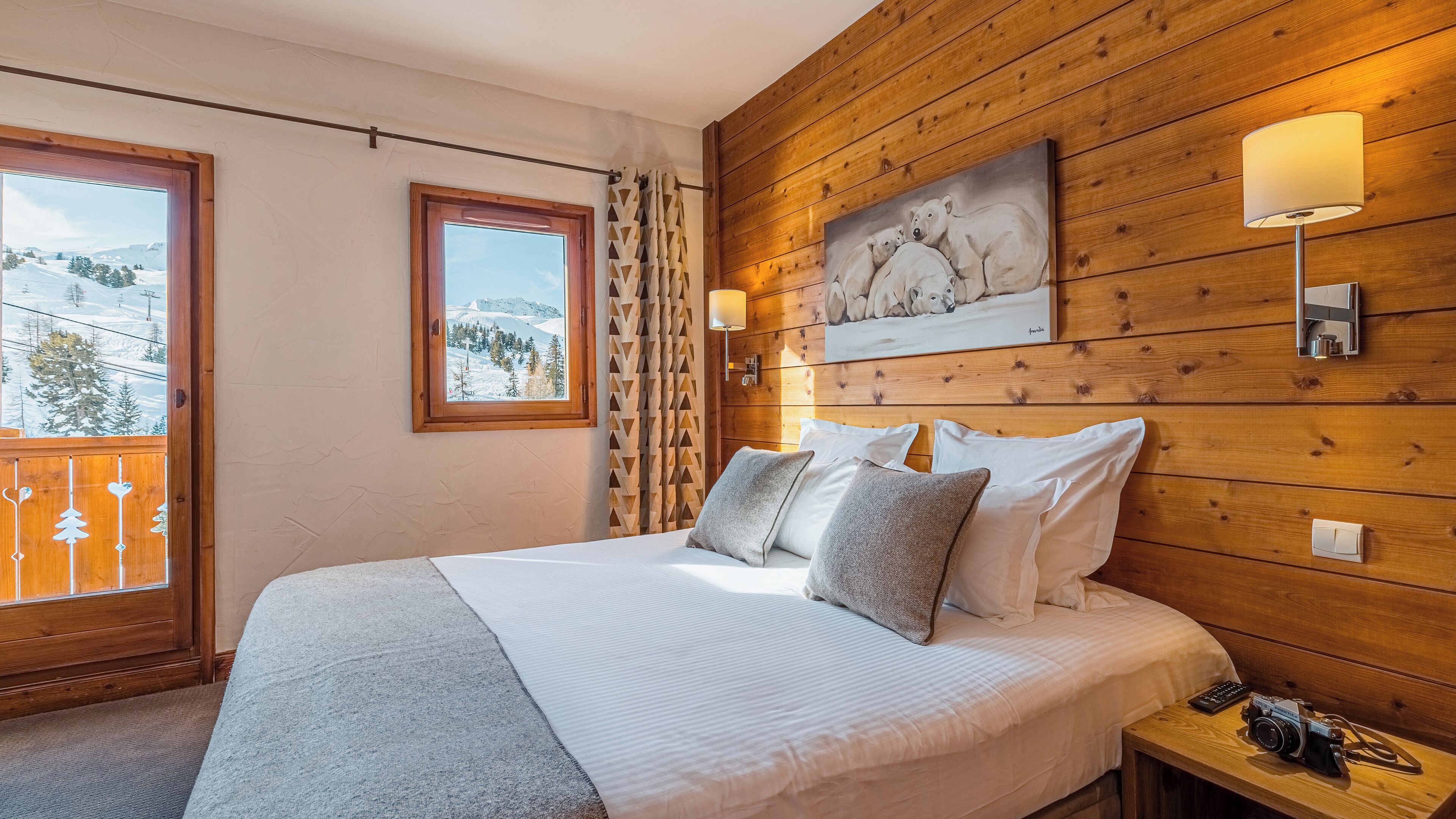 Un dormitorio con cabecera de madera, una cama con ropa blanca y un cuadro de osos polares, y vista a un paisaje nevado a través de una ventana.