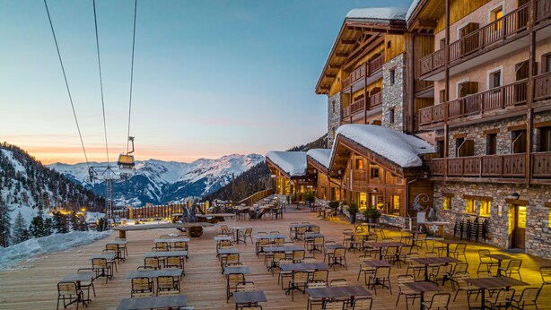 Un espace extérieur aménagé en bois, avec des tables et des chaises, offrant une vue sur une chaîne de montagnes enneigées.