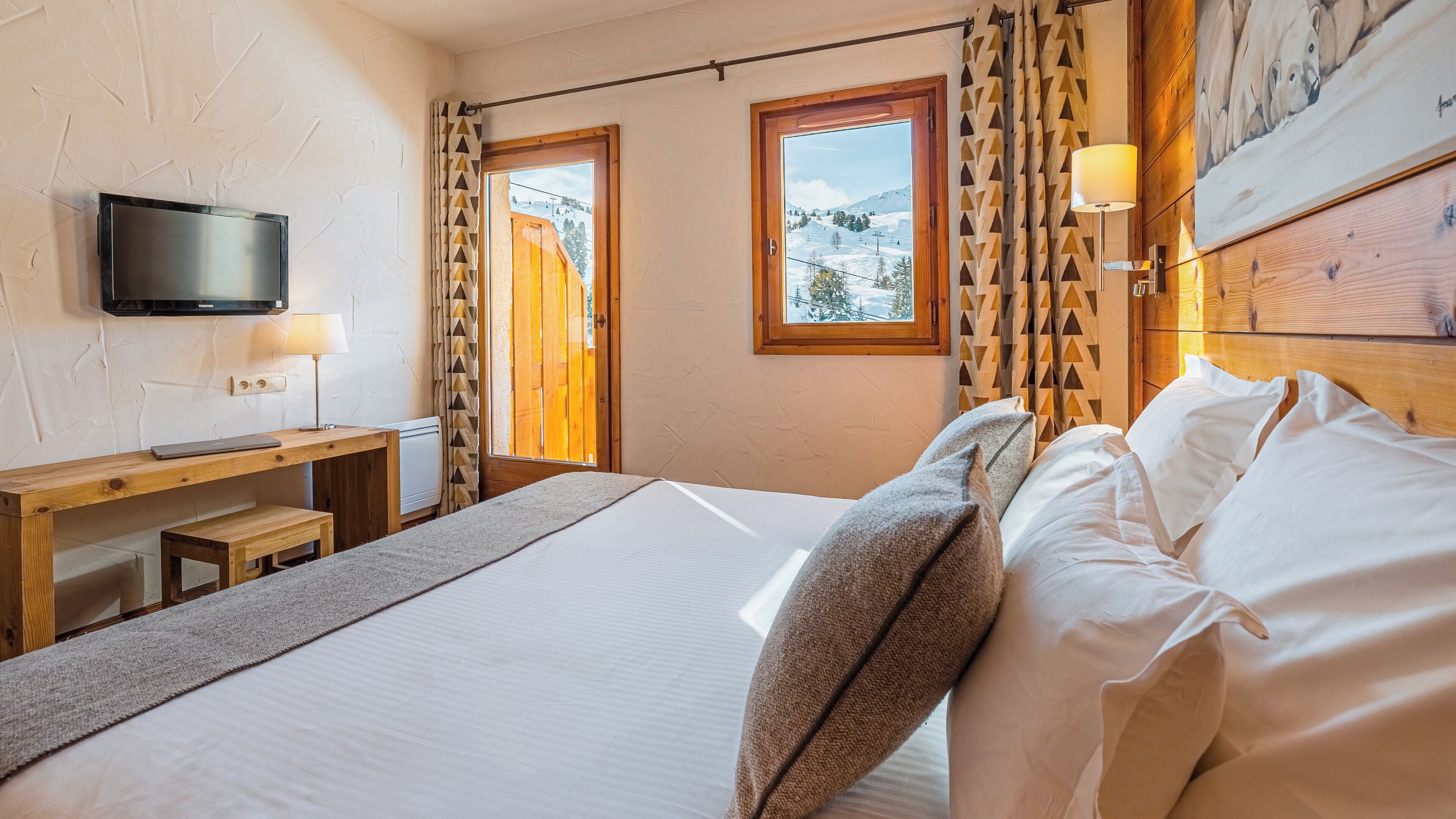 A hotel room with a bed, a desk with a laptop, a TV, and a window with a mountain view.
