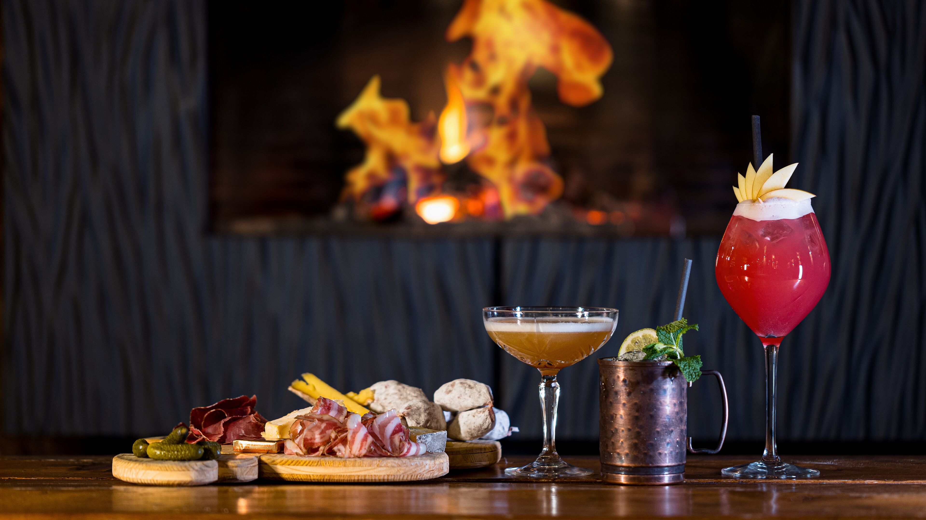 Un bar con una variedad de quesos, embutidos y una selección de bebidas, incluyendo un cóctel y una bebida roja con guarnición.