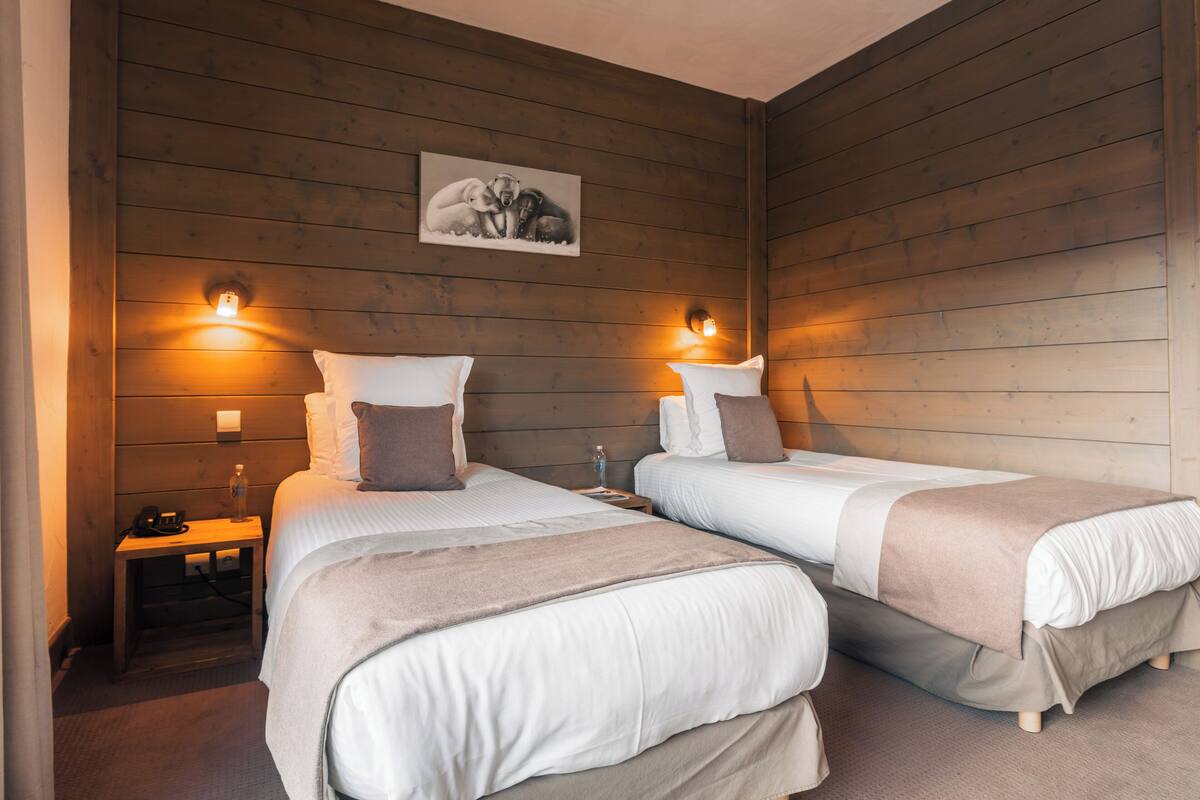A room with two beds, wooden walls, a painting, wall-mounted lights, and a bedside table with a lamp.