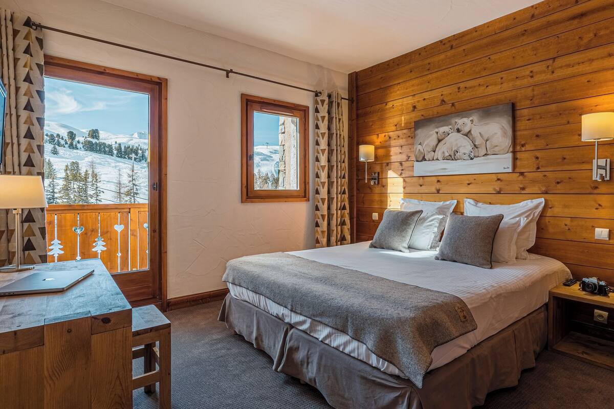 A bedroom with a bed, bedside lamps, a painting, a desk with a lamp, and a view of a snowy landscape.
