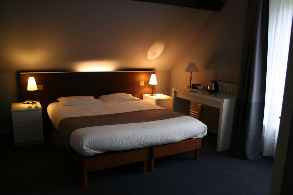 A hotel room with a large bed, two bedside lamps, a nightstand, a desk, and a radiator.
