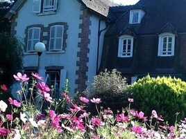 Uma casa de dois andares com fachada branca e telhado escuro, rodeada por um jardim cheio de flores cor-de-rosa.