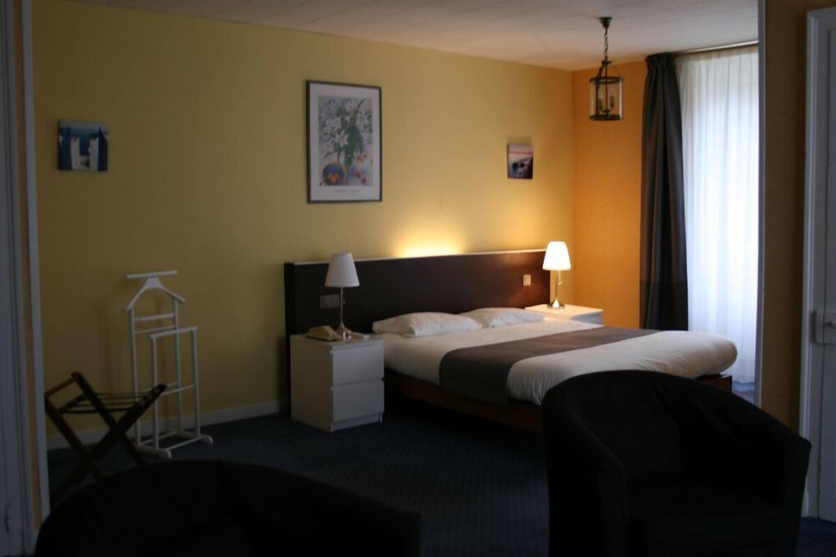 A hotel room with a bed, two bedside lamps, a nightstand, a chair, a coat rack, and a painting on the wall.