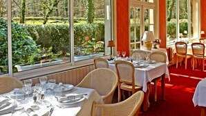 A restaurant with tables set for dining, large windows with a view of greenery, and wicker chairs. - Hotel Le Tronçais (Saint-Bonnet-Troncais)