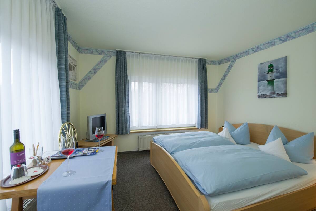 Quarto de hotel com uma cama, uma escrivaninha com computador, uma janela com cortinas, um quadro na parede e um tabuleiro com garrafa e copos.