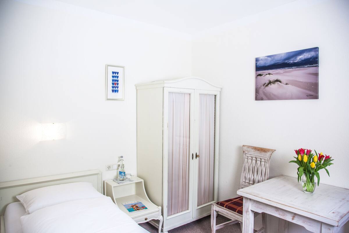 Um quarto com cama, guarda-roupa, mesa de cabeceira com uma garrafa e uma revista, pequena mesa com um vaso de flores, e um quadro na parede.
