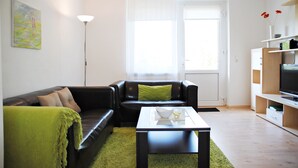 Een moderne woonkamer met een zwarte leren bank, een groene plaid, een witte salontafel en een groot raam met dunne gordijnen.