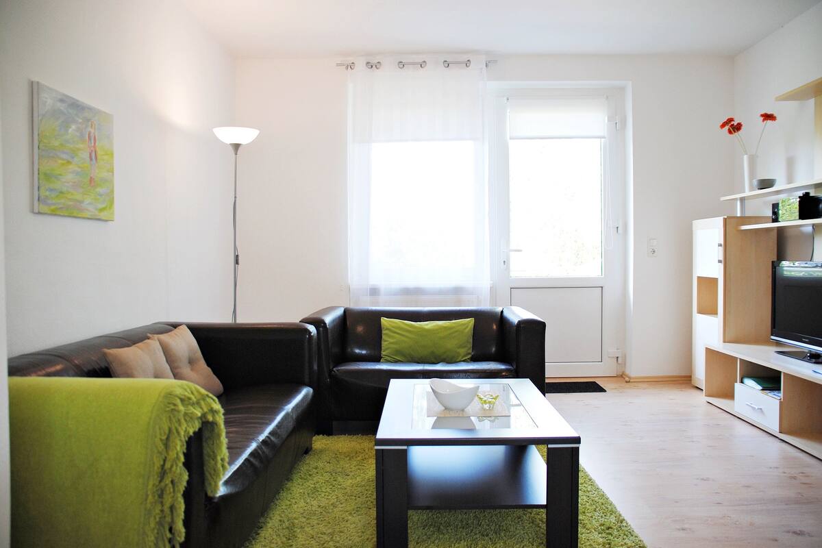 Ein modernes Wohnzimmer mit einem schwarzen Ledersofa, einer grünen Decke, einem weißen Couchtisch und einem großen Fenster mit transparenten Vorhängen.